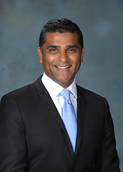 A man standing in front of a background with a microphone, wearing a dark suit and tie, smiling and looking directly at the camera.