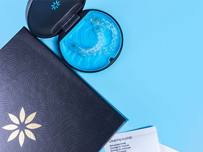 The image shows a closed blue box with a white lid, containing a transparent plastic tray filled with several small, round, light-blue crystals, placed on a surface alongside an open black book with a gold emblem on its cover and a pair of clear glasses with a stylized design on the arms.