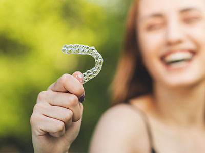 The image shows a person holding up a clear plastic dental retainer with a smile on their face, against a blurred outdoor background.