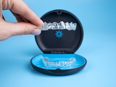 An image showing a person s hand holding an open clear plastic dental retainer case containing a blue and white dental retainer, with a close-up view of the retainer inside.