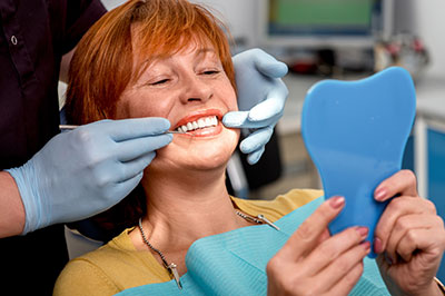 The image shows a woman sitting in a dental chair with her mouth open, receiving dental care from a professional who is using a blue dental tool on her teeth.