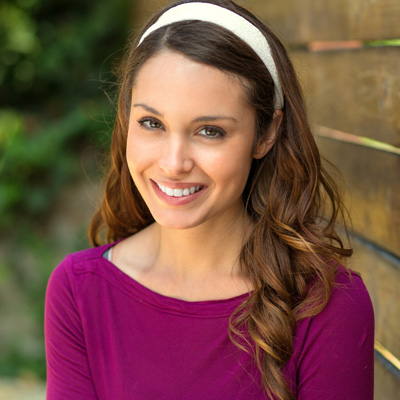 A young woman with long brown hair, wearing a purple top, stands against a wooden fence with a radiant smile on her face.